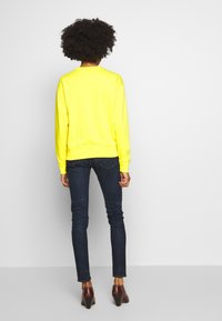 Jasně žlutá mikina s kulatým výstřihem, žebrovanými manžetami a lemem, doplněná tmavě modrými úzkými džínami a hnědými botami na podpatku.