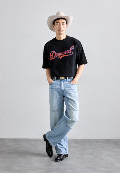 Homem com um chapéu de cowboy branco, t-shirt preta da Dsquared2, jeans de perna larga azul claro, cinto preto e botas pretas, de pé com as mãos nos bolsos.