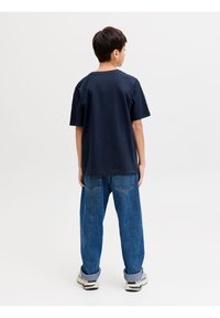 Αγόρι που στέκεται με το πρόσωπο προς τα πίσω φορώντας μπλε μπλουζάκι ναυτικού χρώματος, μπλε τζιν με σηκωμένα ρεβέρ και άσπρα αθλητικά παπούτσια, σε απλό λευκό φόντο.