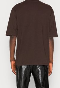 T-shirt en coton marron à manches courtes avec une coupe décontractée, présentant un col rond et une texture lisse, porté avec un pantalon en cuir noir brillant.