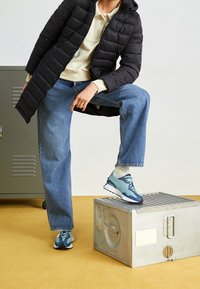 Zapatillas de deporte azul claro y marino con una superficie texturizada, combinadas con jeans azul claro y un abrigo acolchado negro. Caja de metal en el fondo.