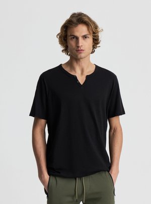 Młody mężczyzna z kręconymi jasnobrązowymi włosami nosi czarną koszulkę typu Henley z krótkim rękawem i oliwkowe spodnie, stoi na jednolitym szarym tle.