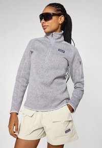 Grijze fleece pullover met een half-rits kraag, voorzien van een Patagonia-logo. Gecombineerd met lichtbeige cargoshorts met meerdere zakken.