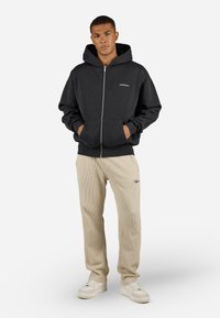 Sort zip-up hættetrøje med snoretræk, forlommer og broderet logo; parret med beige ribbede bukser og hvide sneakers.