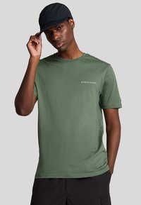 Grøn bomulds t-shirt med korte ærmer, rund hals og lille hvid tekstlogo på brystet, parret med en sort cap.