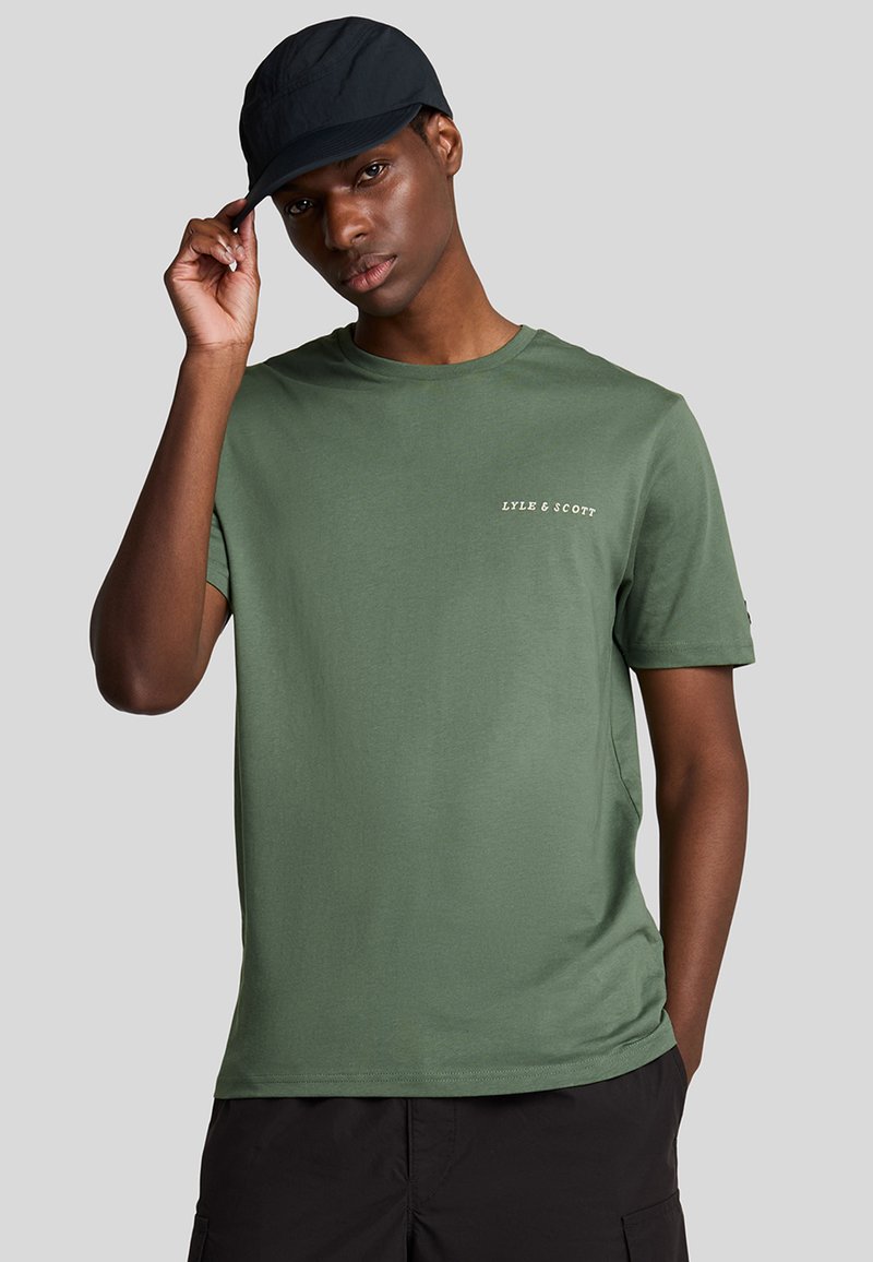 Grøn bomulds t-shirt med korte ærmer, rund hals og lille hvid tekstlogo på brystet, parret med en sort cap.