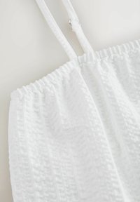 Vêtement blanc texturé sans manches avec bord supérieur élastiqué et bretelles étroites réglables sur fond clair.