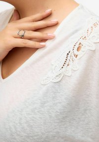 White short-sleeve shirt with a V-neck and delicate lace detailing on the shoulder. Soft, lightweight fabric with a slightly textured finish.