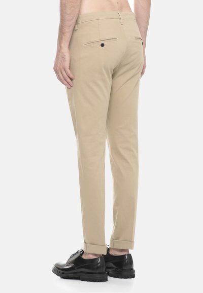 Uomo con pantaloni beige su misura e scarpe in pelle nere, visto di spalle su uno sfondo bianco uniforme.