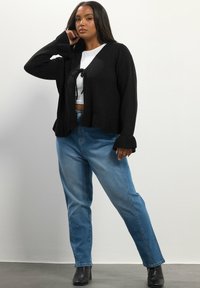 Musta neulevillatakki, jossa on solmittava suljenta, pitkät hihat ja ribattu hihansuu, yhdistettynä vaaleansinisiin suoriin farkkuihin ja mustiin korollisiin saappaisiin.