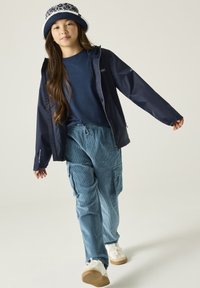 Joven con chaqueta azul marino, camisa azul de manga larga, pantalones cargo de pana azules, zapatillas blancas y un sombrero tipo bucket azul marino con flores caminando hacia adelante.