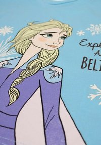 T-shirt bleu avec un graphisme d'un personnage aux longs cheveux blonds, portant une robe violette, entouré de flocons de neige et du texte "Explore As BELIEVE."