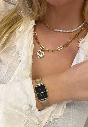 Montre-bracelet à maillon en or avec un cadran noir rectangulaire, superposée à un collier en chaîne à maillons dorés avec un pendentif en forme de cœur et une rangée de perles.