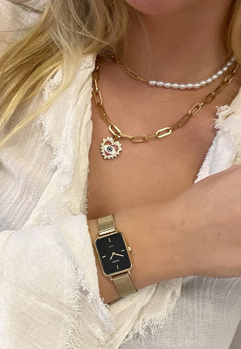 Montre-bracelet à maillon en or avec un cadran noir rectangulaire, superposée à un collier en chaîne à maillons dorés avec un pendentif en forme de cœur et une rangée de perles.