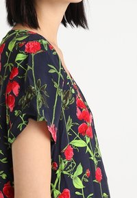 Marinebluse mit kurzen Ärmeln, die ein florales Muster in Rot und Grün aufweist. Der Stoff hat eine glatte Textur und einen lässigen Schnitt.