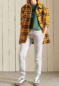 Camisa chaqueta de cuadros amarillos y negros sobre una camiseta verde, combinada con pantalones de chándal grises y zapatillas blancas con acentos verdes y marrones.