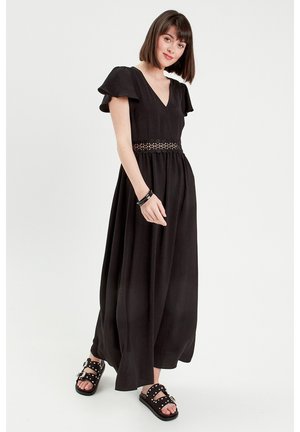Abito maxi nero con maniche corte a campana, scollatura a V e una cintura con motivo in pizzo. Modella abbinato a sandali neri con borchie.