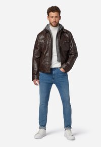 Braune Lederjacke mit einem grauen Kapuzenfutter, ausgestattet mit Klapptaschen und Reißverschlüssen. Getragen über einem weißen Rollkragenpullover und kombiniert mit blauen Jeans und weißen Turnschuhen.