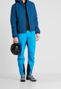 Personne portant une veste bleue et un pantalon bleu vif, tenant un casque noir, debout devant un fond clair uni.