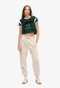 T-shirt vert court avec des accents blancs et l'inscription "CALIFORNIA 48", pantalon de survêtement beige et baskets blanches avec des semelles marron. Un pull en tricot texturé est noué autour des épaules.