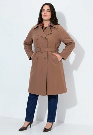 Mujer con un abrigo trench marrón con cinturón sobre jeans oscuros y tacones negros, de pie con las manos en los bolsillos del abrigo frente a un fondo liso.