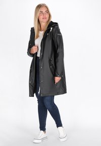 Čierna nepremokavá bunda s kapucňou, predným zipsom, cvokmi a dvomi bočnými vreckami, vrstvená cez bielu košeľu a tmavé džínsy.