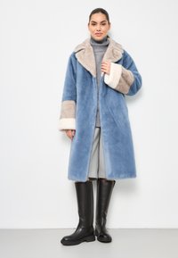 Palton din blană artificială de culoare bleu deschis, cu accente bej și crem, guler larg și mâneci lungi. Purta peste un pulover gri și asortat cu botine negre până la genunchi.