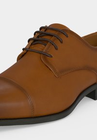 Chaussure habillée en cuir marron avec bout droit et lacets noirs, avec semelle noire et talon bas, vue de côté.
