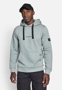 Light teal hoodie with black drawstrings and pocket. Features "REDEFINED REBEL" text and a patch on the sleeve. Soft material, relaxed fit.
