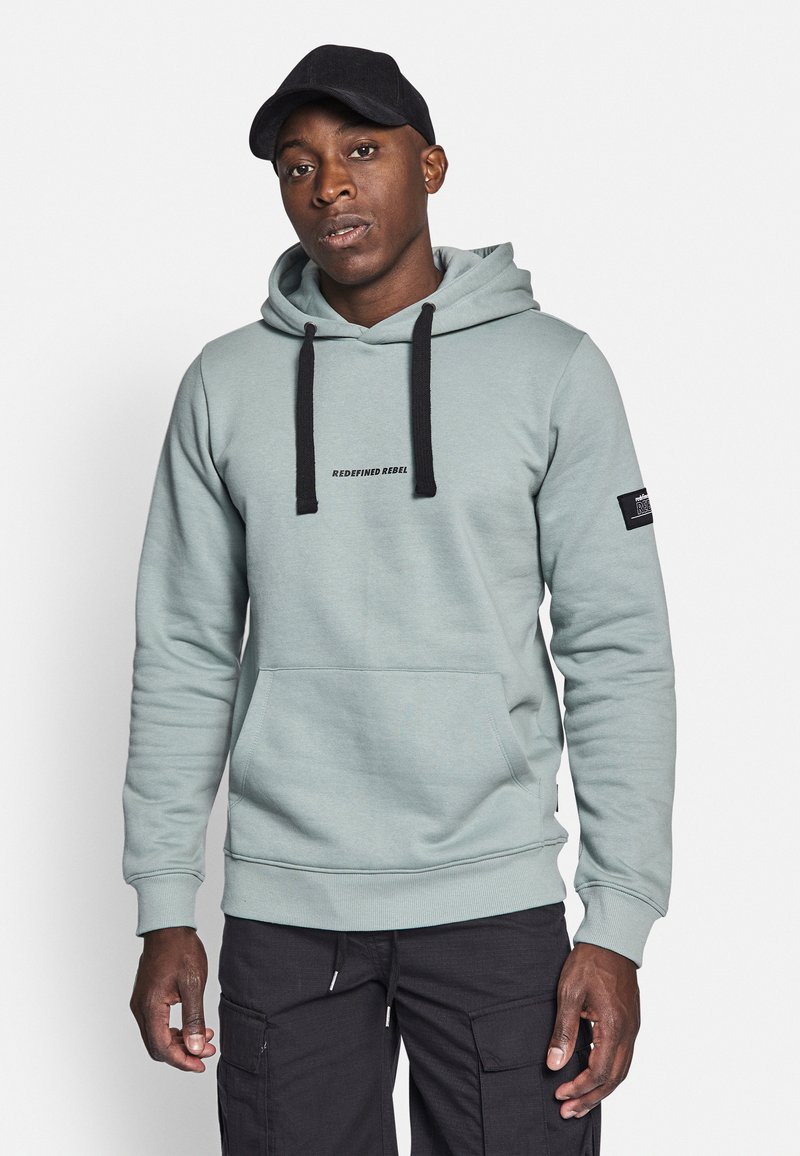 Light teal hoodie with black drawstrings and pocket. Features "REDEFINED REBEL" text and a patch on the sleeve. Soft material, relaxed fit.