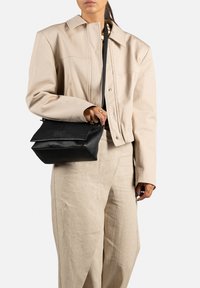 Černá kožená crossbody taška s skládacím designem, texturovaným povrchem a malou značkovou etiketou. Nosit s bledě béžovým sáčkem a kalhotami.