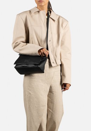 Sac en cuir noir porté en bandoulière avec un design plié, une surface texturée et une petite étiquette de marque. Porté avec une veste et un pantalon beige clair.