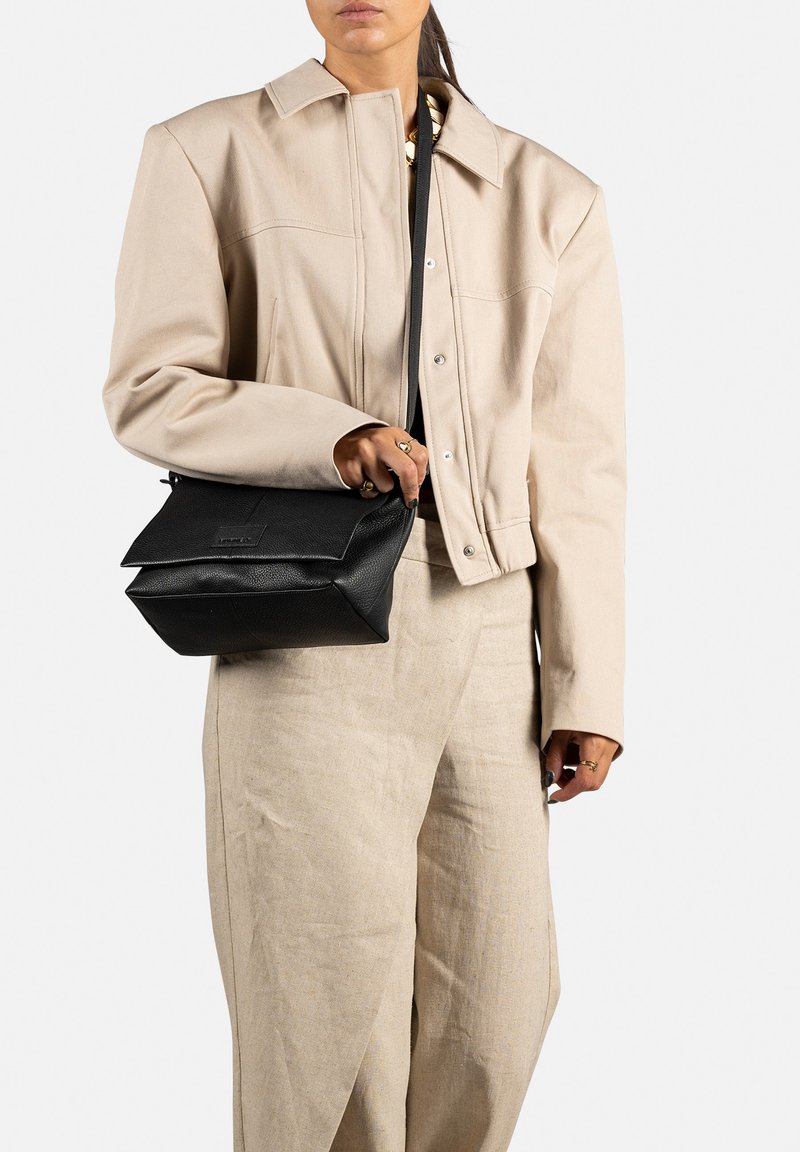 Černá kožená crossbody taška s skládacím designem, texturovaným povrchem a malou značkovou etiketou. Nosit s bledě béžovým sáčkem a kalhotami.