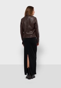 Veste en cuir marron au design slim, portée sur une robe maxi noire avec une fente haute sur la cuisse. Des bottes noires épaisses complètent l'ensemble.