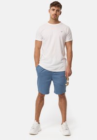 Hvid t-shirt med korte ærmer og blå shorts, holdt med den ene hånd. Hvide sneakers med struktureret sål og solbriller i den anden hånd.