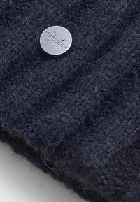 Tmavě námořnická pletená látka s měkkým, texturovaným povrchem a stříbrným kulatým kovovým štítkem vyrytým s nápisem "PEARLWOOD" a dekorativními liniemi.