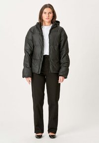 Sort pufferjakke med hætte, der har en quiltet tekstur og frontlynlås. Båret over en hvid skjorte, parret med højtaljede sorte bukser.