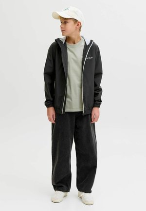 Dječak u bijeloj kapici, crnoj jakni na patentni zatvarač, svijetlozelenoj majici, širokim crnim trapericama i bijelim tenisicama stoji i gleda ulijevo.