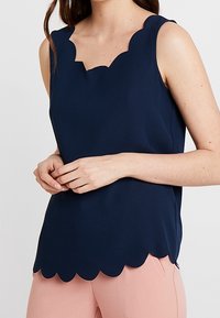 Frau in einem ärmellosen navyblauen Top mit gewellten Kanten, kombiniert mit hellrosa Hosen, die ihre Hände sanft vor sich gefaltet hat.