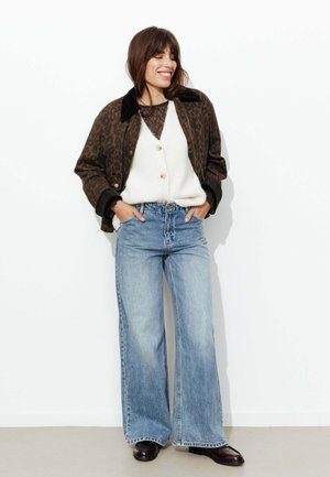 Femme souriante, portant une veste marron à imprimé léopard, un cardigan blanc boutonné, un jean bleu à jambes larges et des chaussures marron foncé, les mains dans les poches.