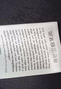 Štítok na pranie zobrazujúci viacjazyčné pokyny na pranie a symboly pre pranie v studenej vode, bez bielidla, sušenie zavesením, žehlenie na nízku teplotu, žiadne chemické čistenie.