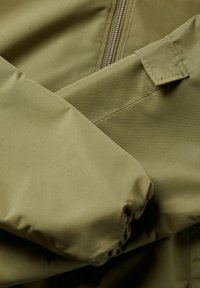 Alkūnės žalia striukė su lygaus, blizgaus paviršiaus, užtrauktuku ir susiraukšlėjusiais rankogaliais. Tiesios linijos su minimaliais akcentais.