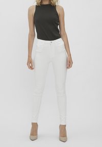 Valkoiset kapeat farkut, jotka on yhdistetty mustaan ribbimäiseen hihattomaan toppiin. Mallilla on päällään beigeit korkokengät, jotka korostavat istuvaa muotoilua.