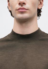 Ung mann iført en brun høyhalset genser, viser nedre del av ansiktet, nakken og øvre bryst mot en enkel hvit bakgrunn.