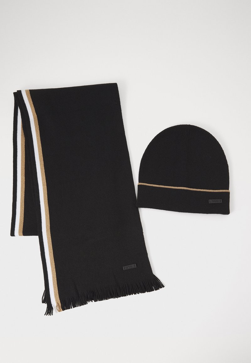 Sort strikket hue med en gylden accent og matchende sort tørklæde, der har hvide og gyldne striber samt frynser i enderne. Begge dele har mærketags.