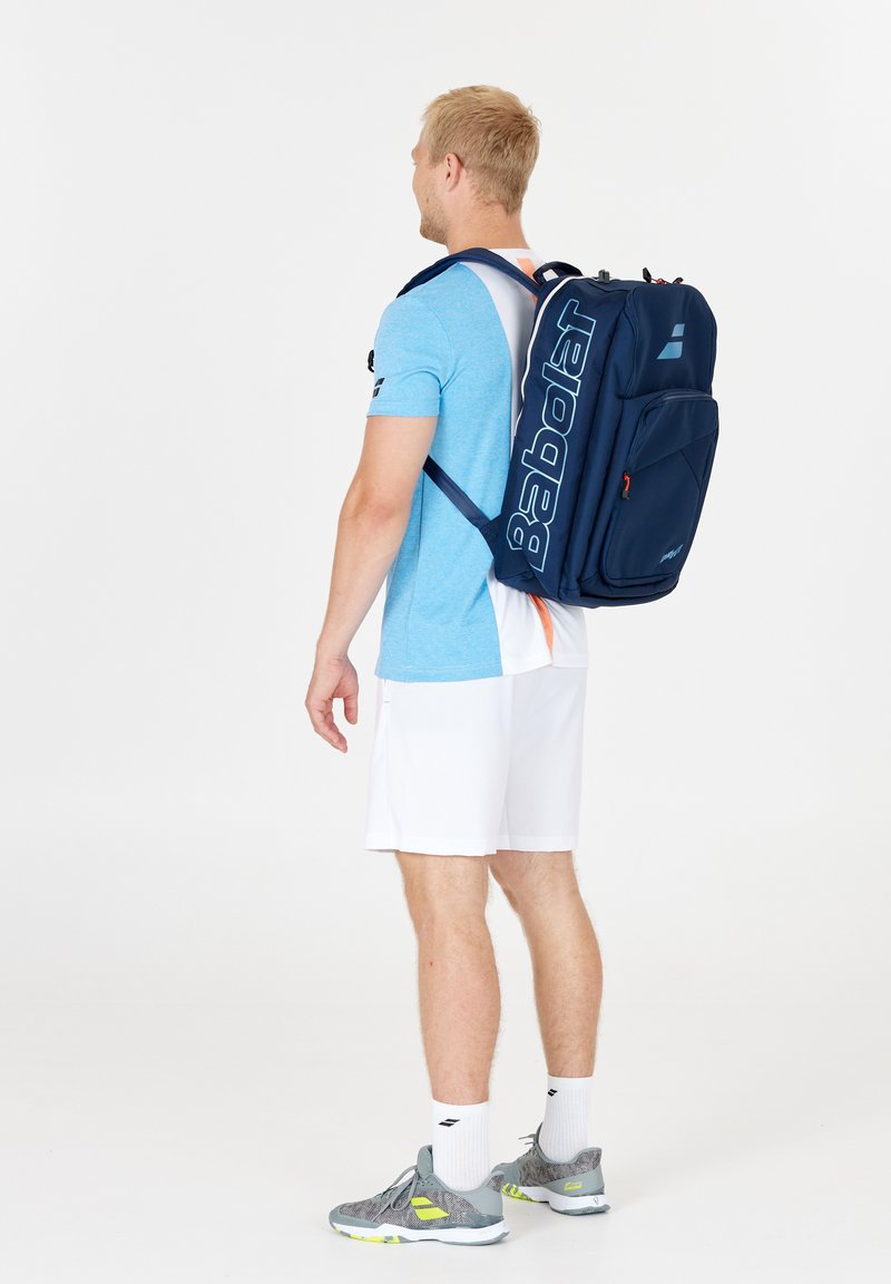 Donkerblauwe rugzak met groot wit "Babolat"-logo, ergonomisch ontwerp, gevoerde banden en voorvak. Draagt door een persoon in lichtblauw shirt en witte korte broek.