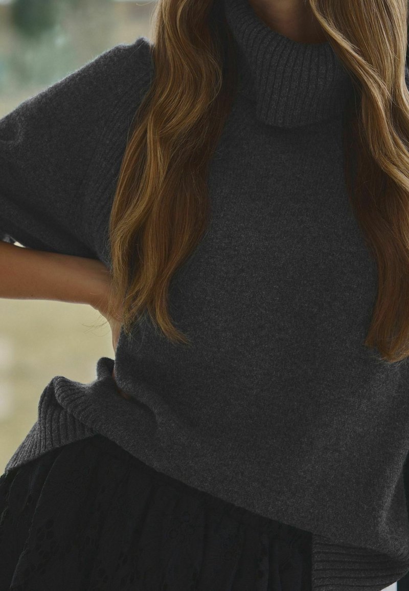 Dunkelgrauer, gestrickter Rollkragenpullover mit weitem, geripptem Kragen und kurzen Ärmeln. Akzente eines strukturierten, plissierten schwarzen Rocks.