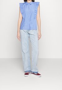 Blusa senza maniche azzurro chiaro con righe verticali, dettagli a volant sulle spalle, chiusura frontale con bottoni, abbinata a jeans chiari e sneakers rosse.