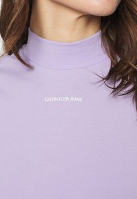Fialový rolák vyrobený z měkké látky s hladkým povrchem, s logem "CALVIN KLEIN JEANS" v bílé barvě na hrudi.