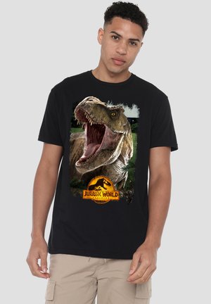 Junger Mann trägt schwarzes T-Shirt mit großem brüllendem Tyrannosaurus Rex und Jurassic World Logo vorne, beige Cargohose.